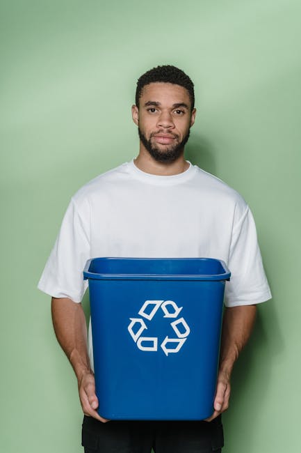 A sanitation worker dressed in a green uniform with orange reflective stripes stands beside an open rear compartment of a waste collection truck, which is parked on a residential street. The truck has
