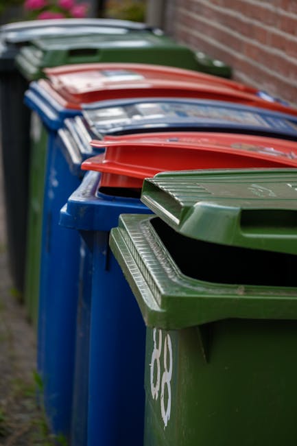 A row of large, rectangular wheelie bins lined up along a pavement beside a brick wall, featuring various coloured lids including green, red, and blue. The green bin in the foreground has a textured, 