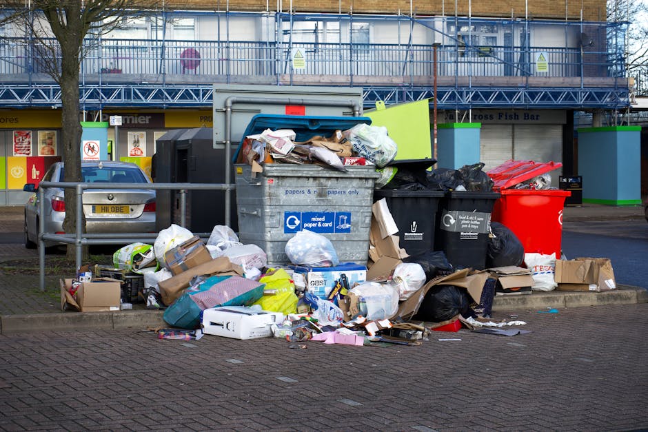 A row of large, rectangular wheelie bins lined up along a pavement beside a brick wall, featuring various coloured lids including green, red, and blue. The green bin in the foreground has a textured, 
